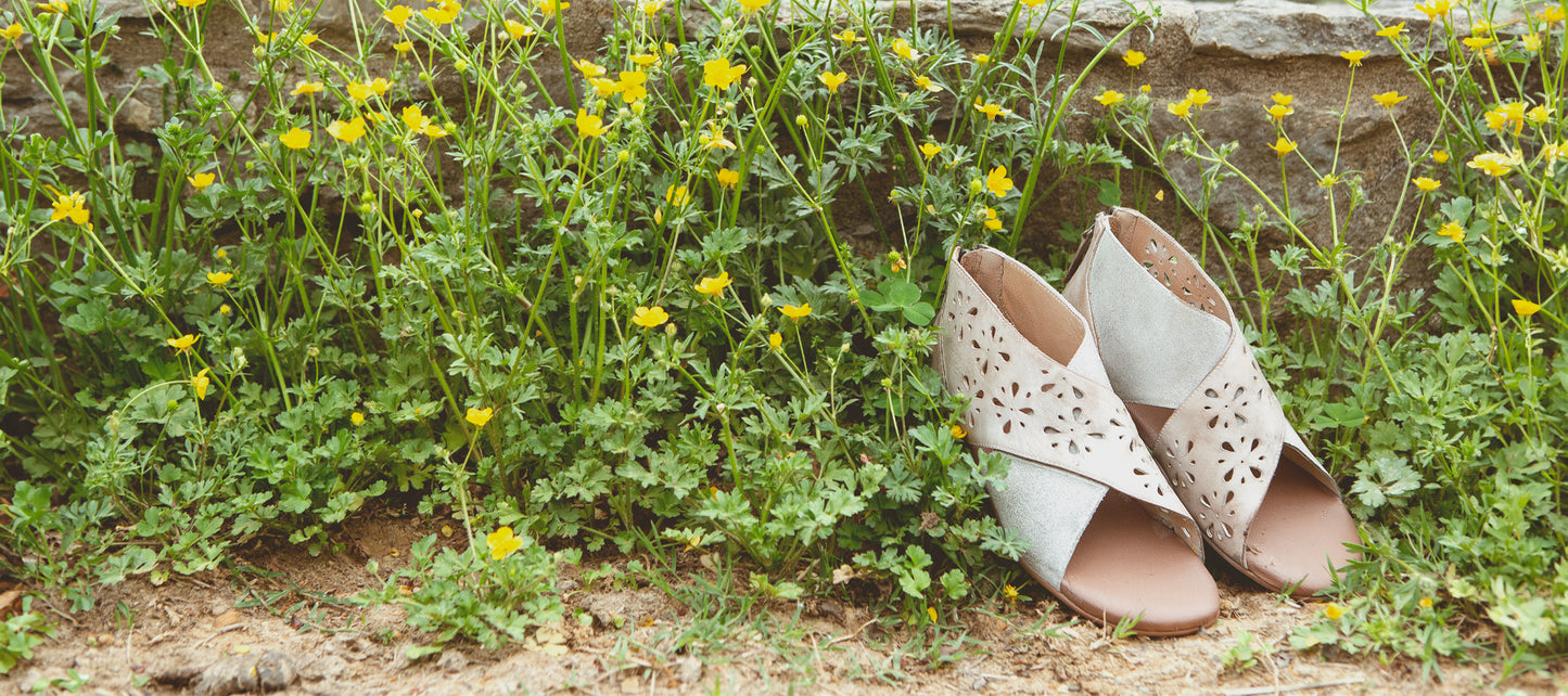 A pair of beige sandals with cut-out details placed on the ground next to a stone wall surrounded by yellow wildflowers.