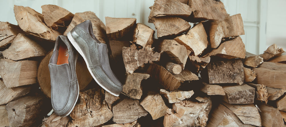 A pair of gray slip-on shoes placed on top of a pile of firewood against a wooden wall.