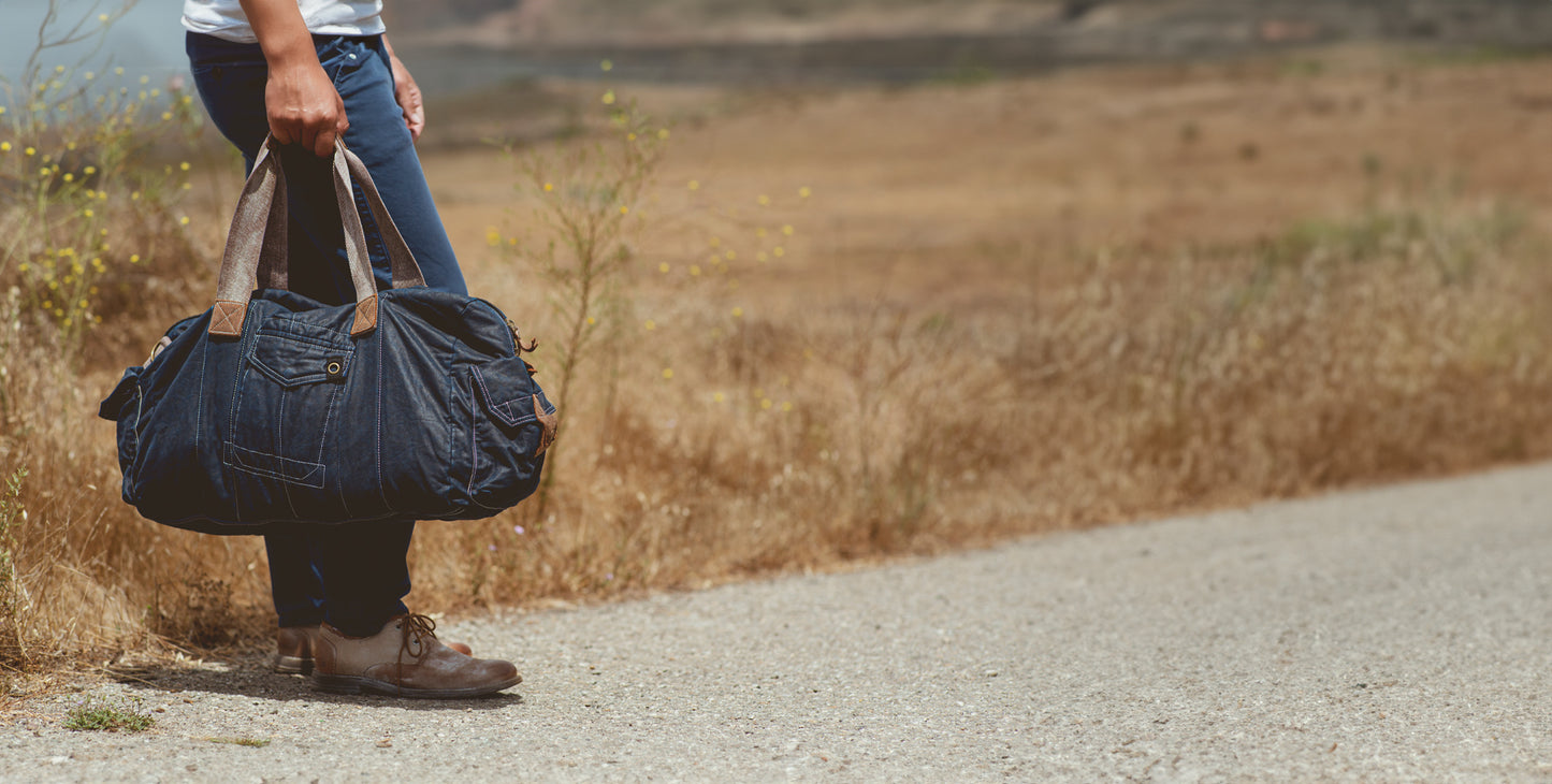 Person holding a large duffel bag standing beside a road with dry grass and a blurred background.