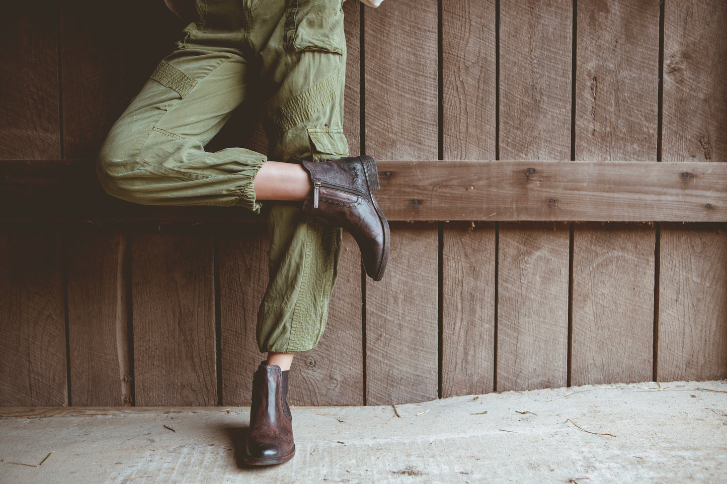 Person wearing green cargo pants and brown boots, leaning against a wooden wall with legs crossed.