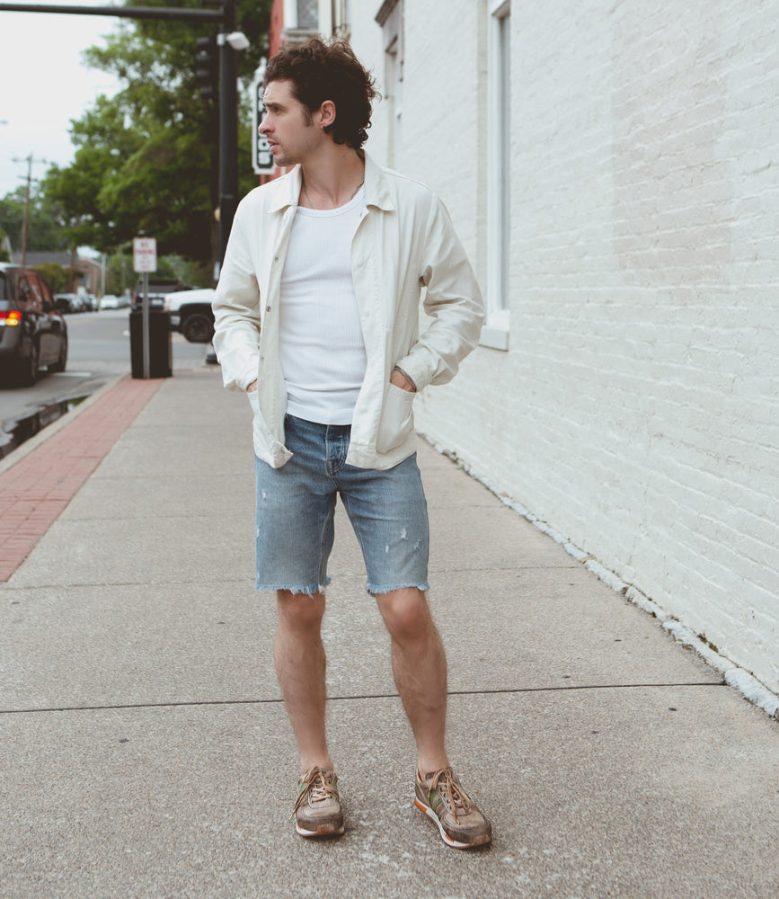 
                  
                    A person wearing a light jacket over a white shirt featuring contrasting green stripes, denim shorts, and Roan's retro-inspired Alatar leather sneakers stands on a sidewalk looking to their left. Brick buildings and parked cars are in the background.
                  
                