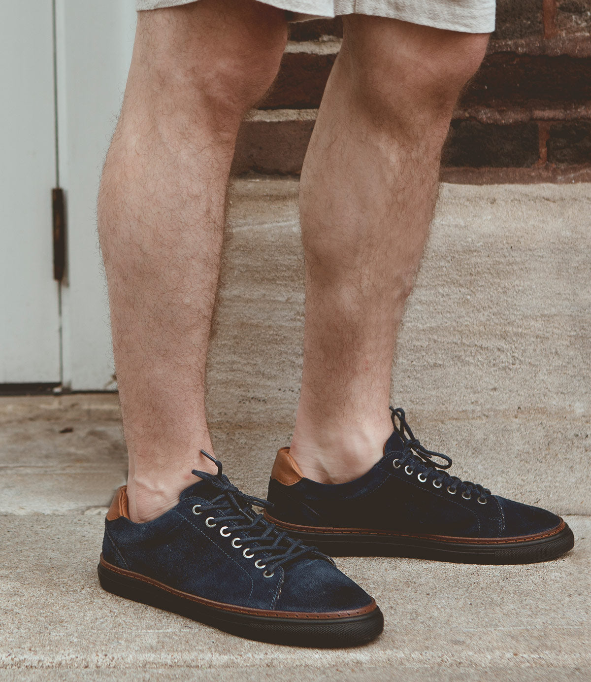 
                  
                    A person wearing versatile blue suede Roan Albright sneakers with brown accents and beige shorts stands on concrete steps near a brick wall.
                  
                