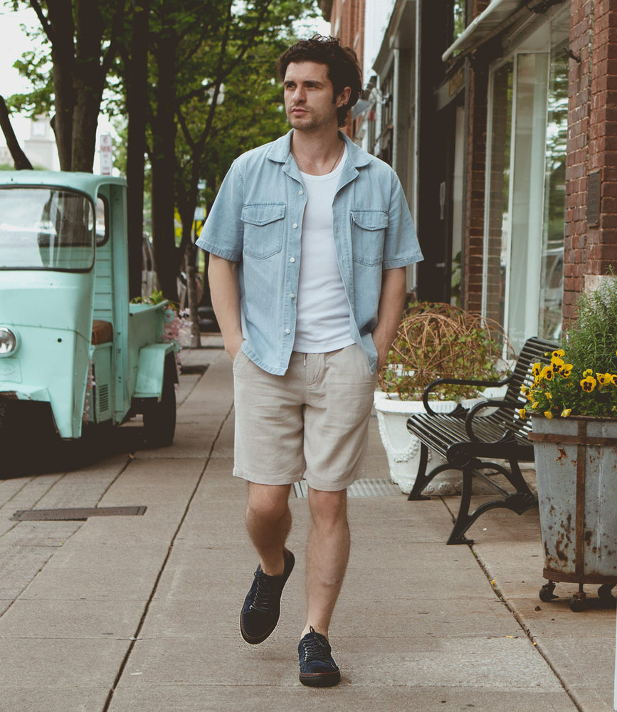 
                  
                    A man in a pale blue short-sleeved shirt, white t-shirt, and beige shorts walks on a sidewalk beside buildings and potted plants, sporting Roan's Albright footwear that perfectly complements his casual style.
                  
                