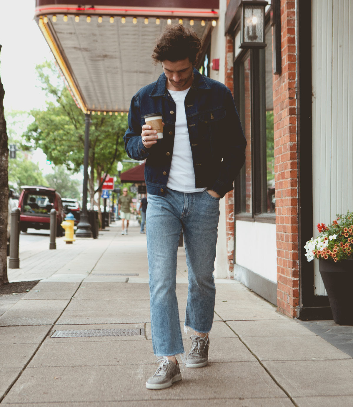 
                  
                    A man in a denim jacket, white shirt, and jeans walks down a sidewalk holding a coffee cup. There are buildings, a planter with flowers, and trees in the background. His Roan Attitude shoes, with real rubber outsole and suede leather finish, complete the look—making him a walking handmade masterpiece.
                  
                
