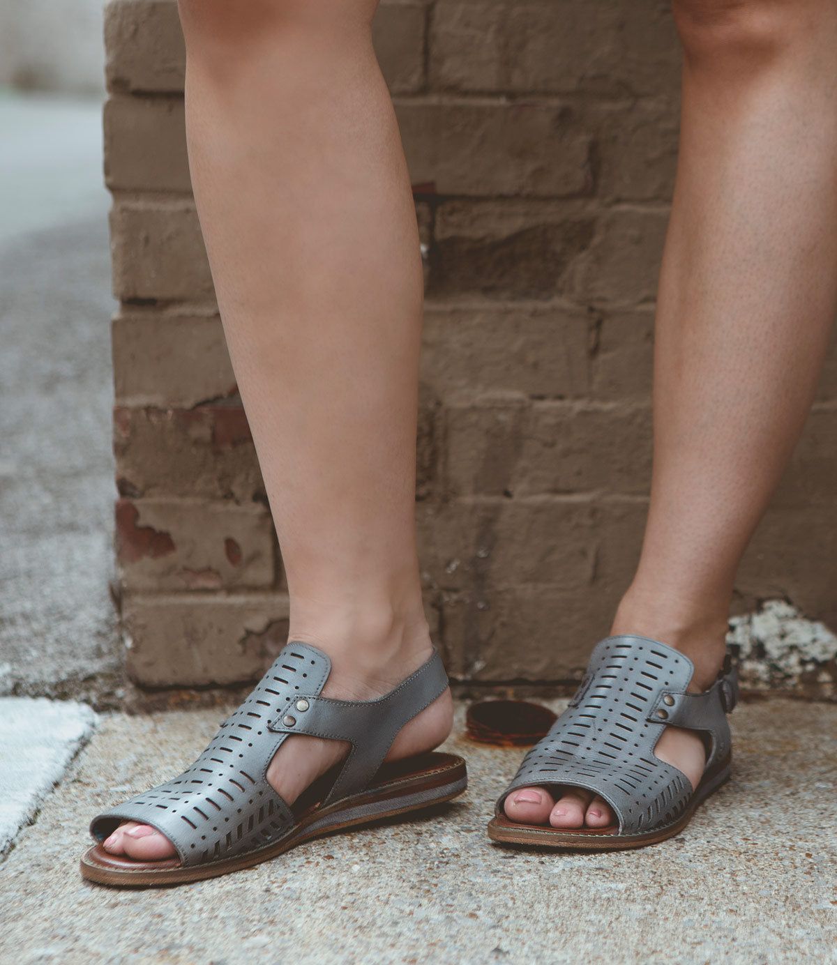 A person wearing gray, open-toed Roan Ballad II sandals with cut-out designs and an adjustable ankle closure stands on a concrete surface near a brick wall.