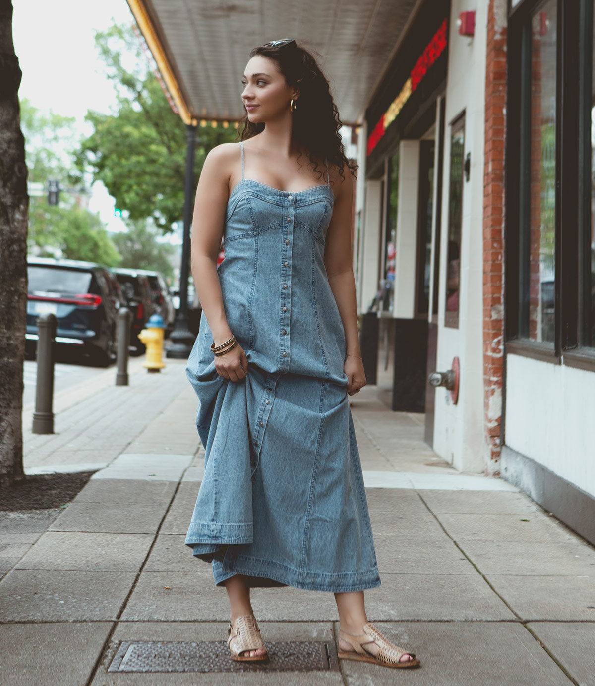 
                  
                    A woman in a strapless denim dress stands on a city sidewalk, looking to the side. She is wearing Roan Ballad II sandals with adjustable ankle closures, and the street behind her features storefronts and parked cars.
                  
                