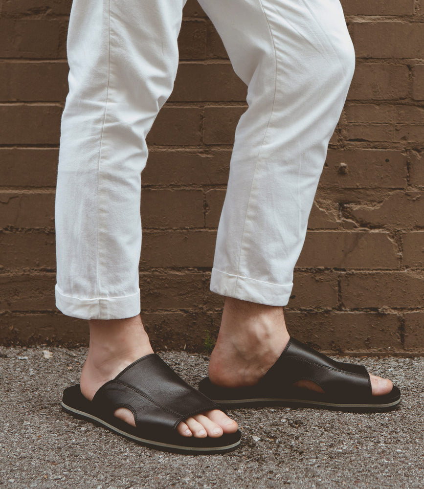 Person wearing white cuffed pants and Roan Breakfast versatile classic slip-on sandals stands outside against a brown brick wall.