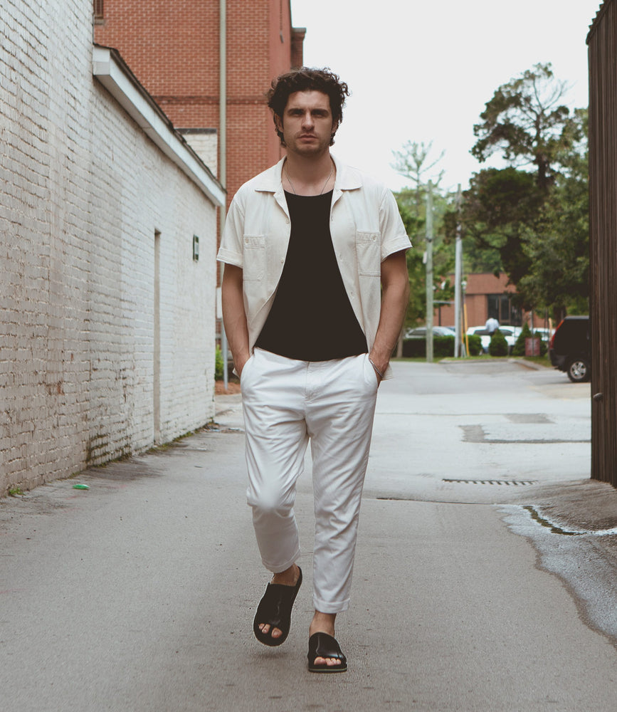 
                  
                    A man walks down an alleyway wearing a light-colored open shirt over a black t-shirt, white pants, and Roan Breakfast slip-on sandals. He steps on a damp pavement bordered by brick walls and greenery.
                  
                