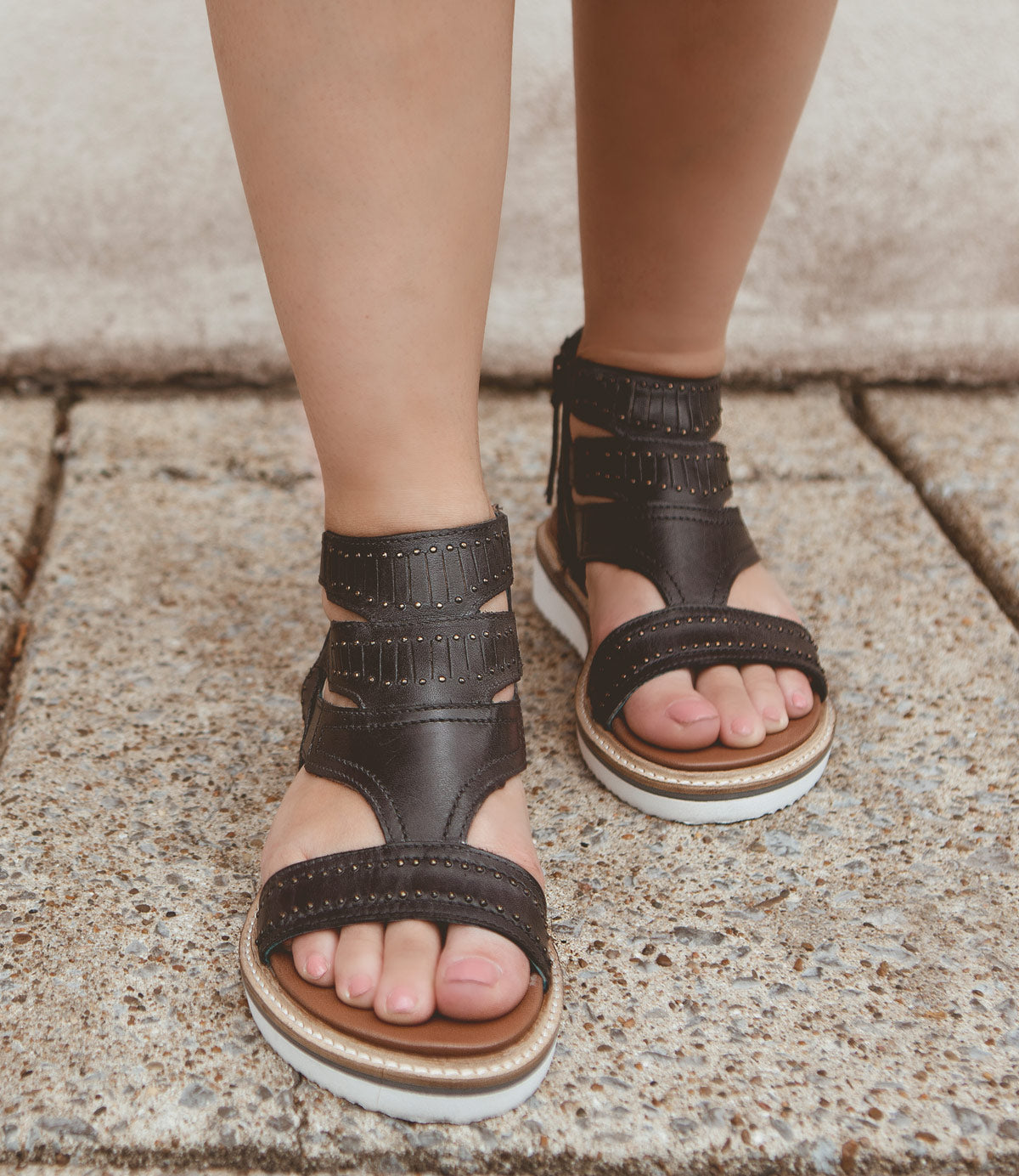
                  
                    A person wearing Roan Carlita II black strappy sandals with white soles, standing on a concrete surface, enjoys cushioned comfort.
                  
                