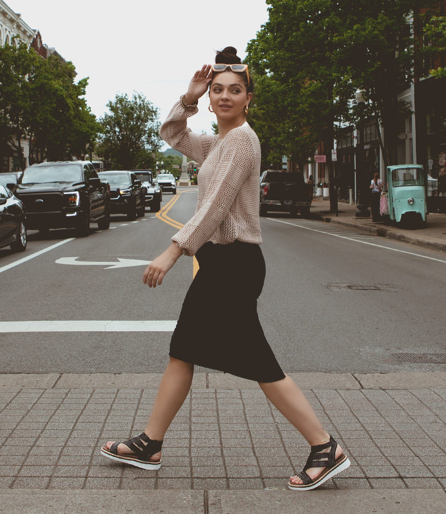 
                  
                    A person in a beige sweater and black skirt walks across a street, holding sunglasses. A small green vehicle and trees are visible in the background, while their shoes feature the cushioned comfort of Roan Carlita II for every step.
                  
                