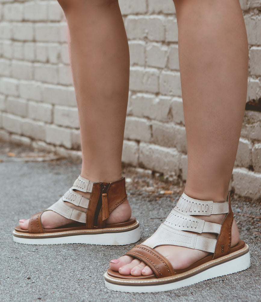 A person wearing Roan Carlita II brown and white strapped sandals with white soles, featuring full-grain leather for durability, stands on a sidewalk next to a brick wall.