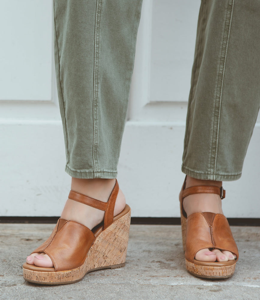 
                  
                    Close-up of a person's feet wearing brown leather wedge heels and olive green pants, standing on a concrete surface in front of a white door. The **Deduction** full-grain leather sandals by **Roan** offer medium coverage, effortlessly blending style and comfort.
                  
                