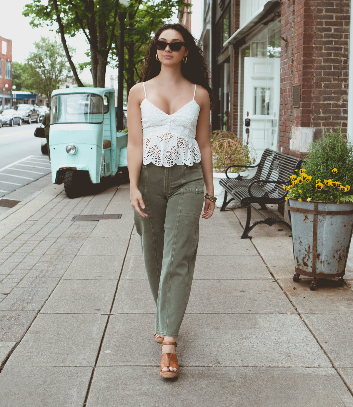 
                  
                    A woman wearing a white lace top and green pants walks down a sidewalk. She is wearing sunglasses and Roan Deduction wedge heels. A vintage turquoise vehicle and potted flowers are in the background.
                  
                