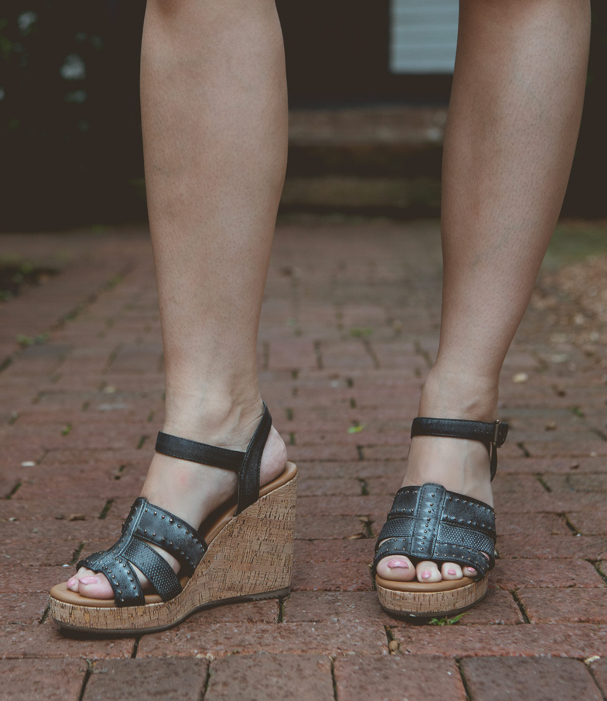 A person wearing Roan Different black, strappy wedge sandals with a leather footbed and wooden platform heels stands on a brick pavement.