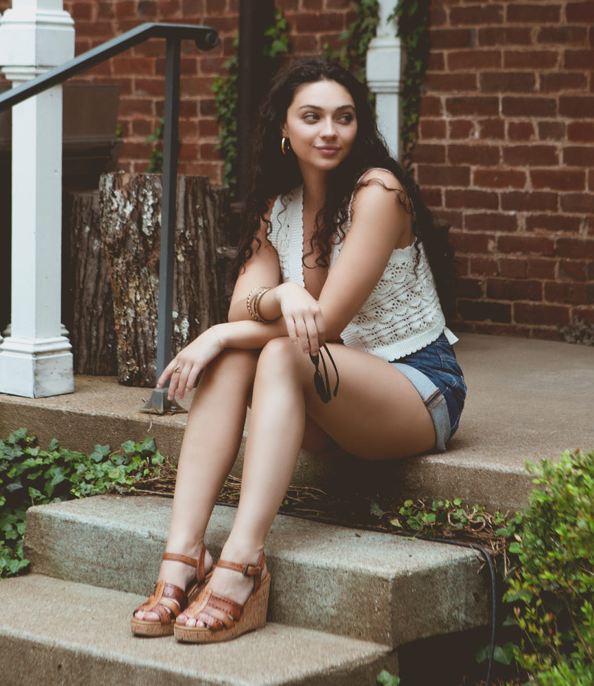 
                  
                    A person with long, dark hair sits on outdoor steps, wearing a white sleeveless top, denim shorts, and Roan Different brown heeled sandals with a padded leather footbed. They are looking to the side, with greenery and a brick wall in the background.
                  
                