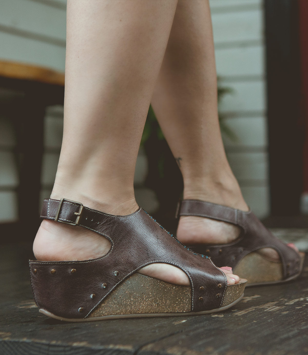 
                  
                    Close-up of a person's feet wearing Roan Fortnight brown full-grain leather wedge sandals with buckle straps and exposed toes. The person is standing on a dark wooden floor, showcasing the adjustable ankle strap for a secure fit.
                  
                
