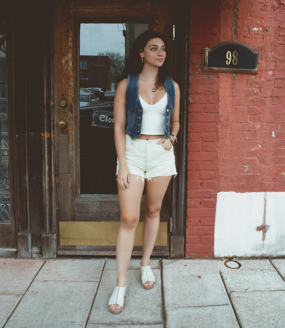 
                  
                    A woman in a denim vest and white shorts stands in front of an old wooden door, with the number 98 displayed on a red brick wall to her right, showcasing Roan Fortnight slip-on platform shoes.
                  
                