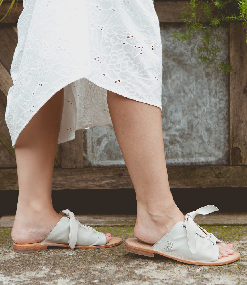 A person wearing Roan Grapevine sandals with adjustable leather straps and a white lace skirt stands on a stone path with greenery and a wooden door in the background.