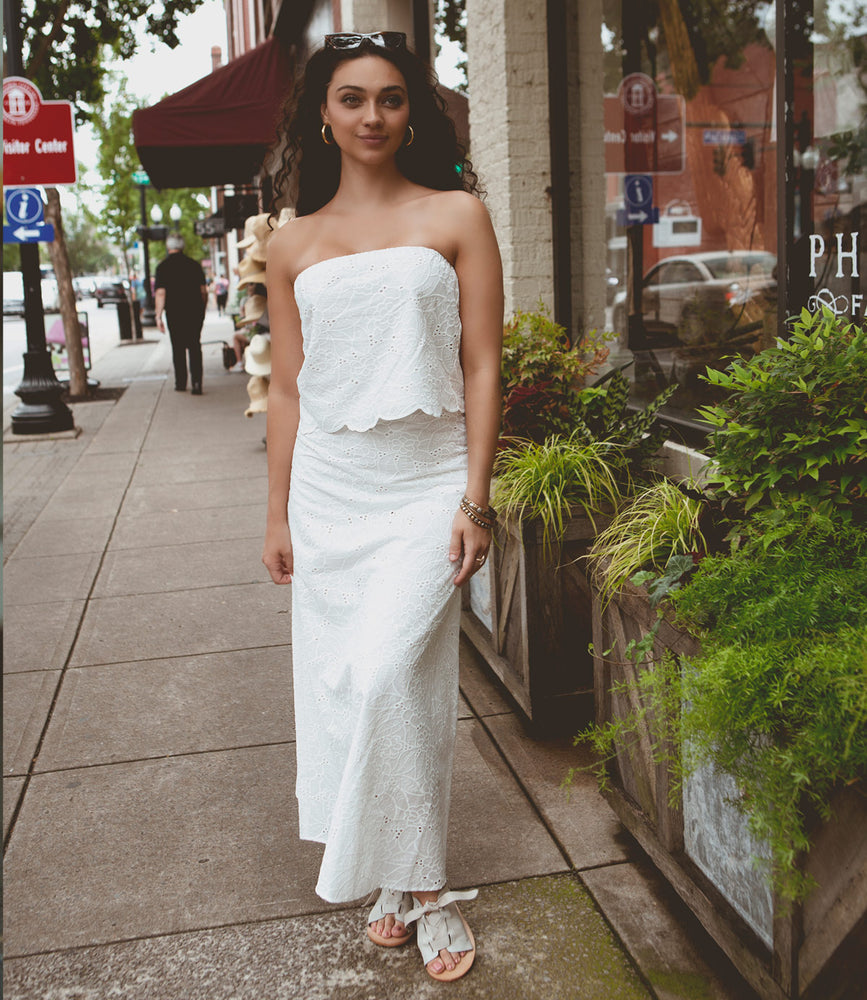 
                  
                    A woman wearing a strapless white dress and Roan Grapevine leather sandals stands on a sidewalk lined with luxury storefronts and lush plants.
                  
                