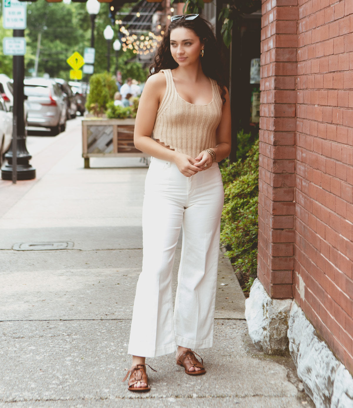 
                  
                    A woman wearing a beige sleeveless top and white pants, complemented by Roan's Grapevine sandals with adjustable leather straps, stands on a sidewalk next to a brick building.
                  
                