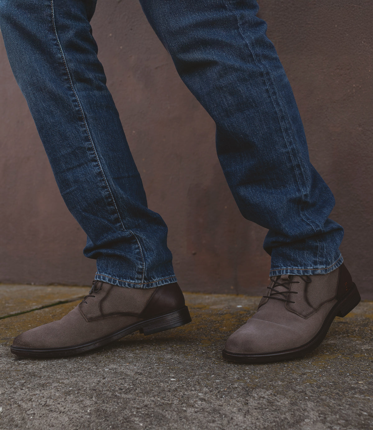 A person wearing the Roan Joyride brown suede lace-up boots and blue jeans stands on a concrete surface against a brown wall.