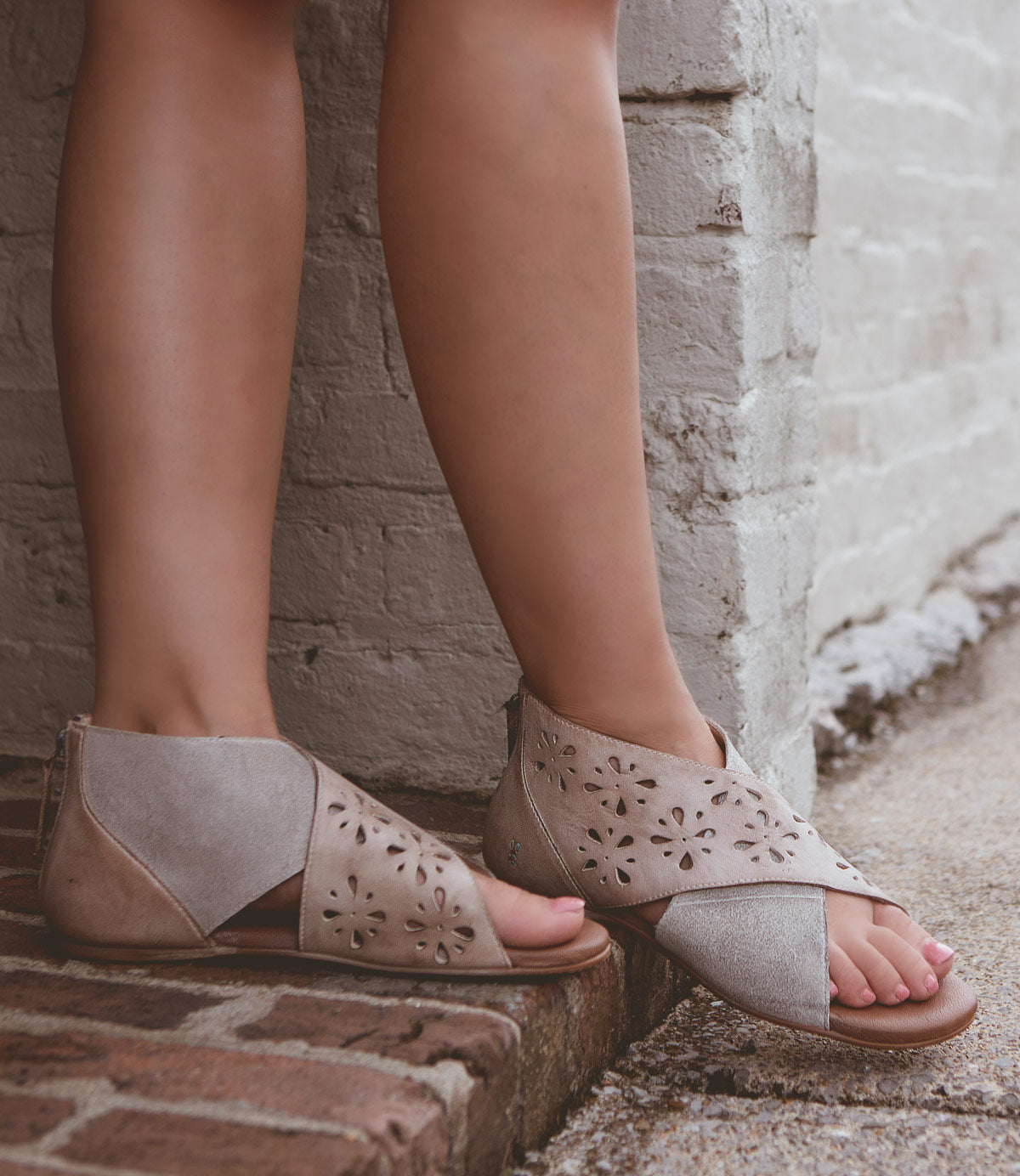 Person wearing Roan Rotation light brown, leather cross-strap sandals with decorative cutouts and a cushioned footbed, standing on a brick step against a white wall.