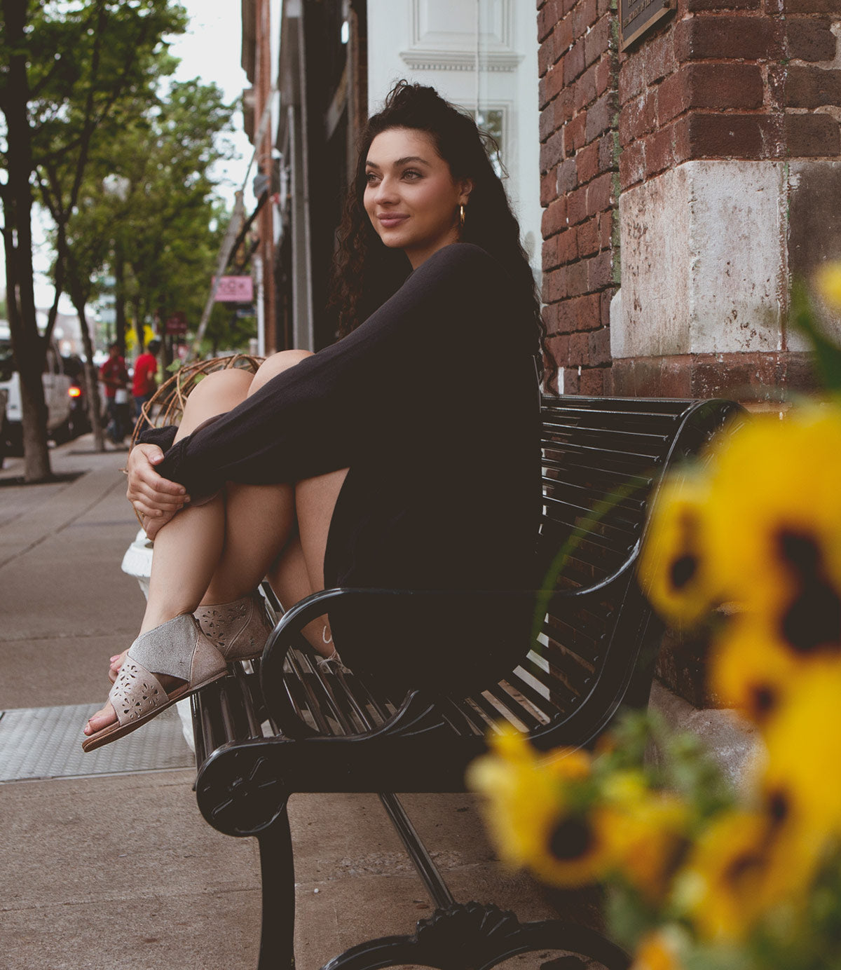 
                  
                    A woman sits on a bench with her arms wrapped around her knees, smiling and looking into the distance. She is wearing a dark sweater and Roan Rotation sandals. Yellow flowers are in the foreground.
                  
                