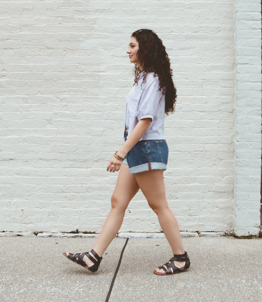
                  
                    A woman with long curly hair walks along a sidewalk in front of a white brick wall, wearing a light shirt, denim shorts, and black leather Royalty sandals by Roan.
                  
                