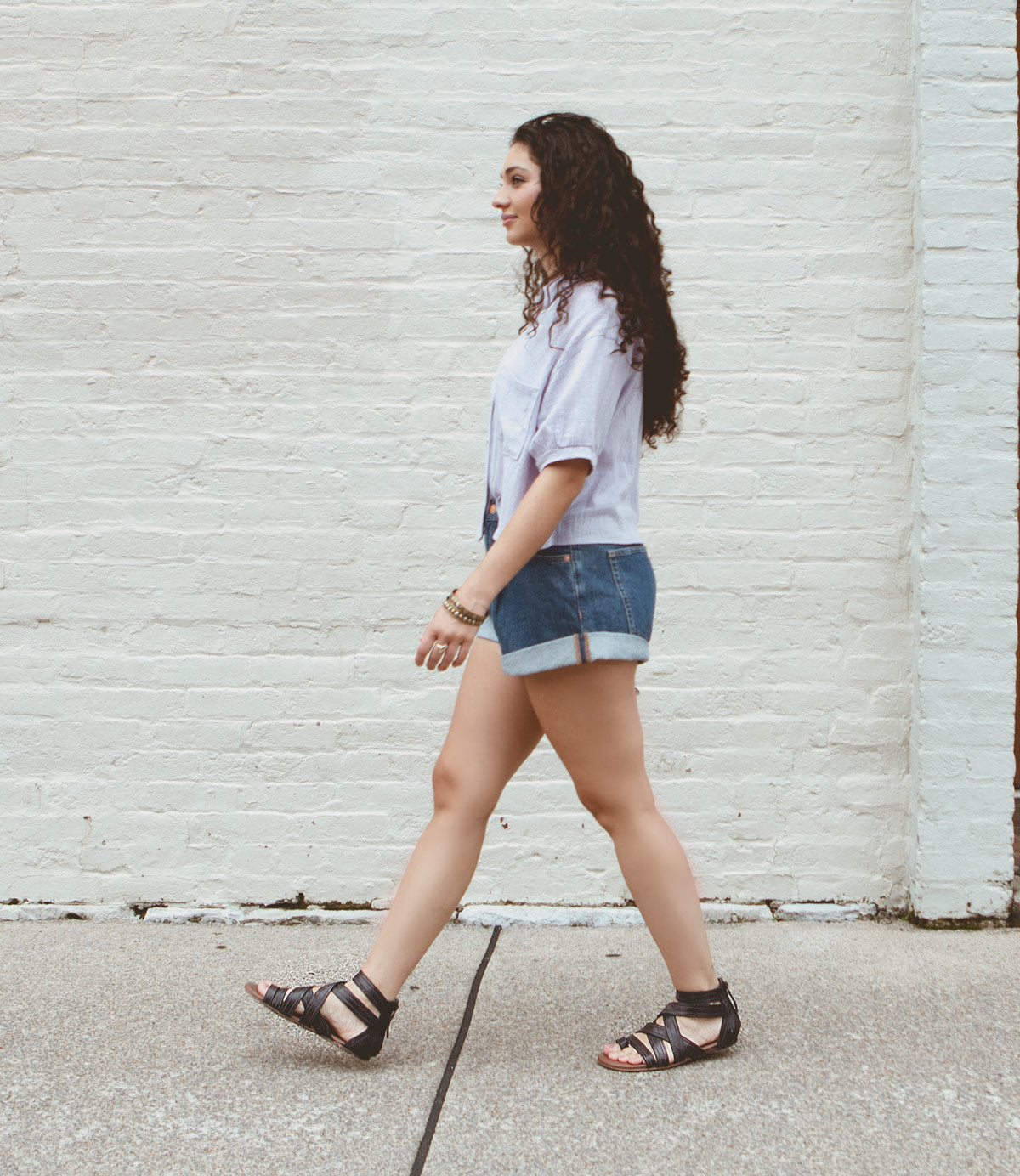 
                  
                    A woman with long curly hair walks along a sidewalk in front of a white brick wall, wearing a light shirt, denim shorts, and black leather Royalty sandals by Roan.
                  
                