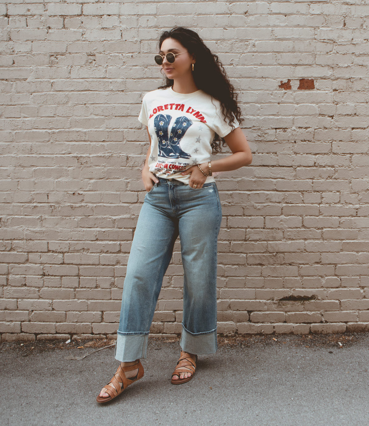 
                  
                    A person with long hair wearing sunglasses, a graphic t-shirt, wide-leg jeans, and Roan Royalty leather sandals with cushioned footbeds stands against a brick wall with hands in pockets.
                  
                