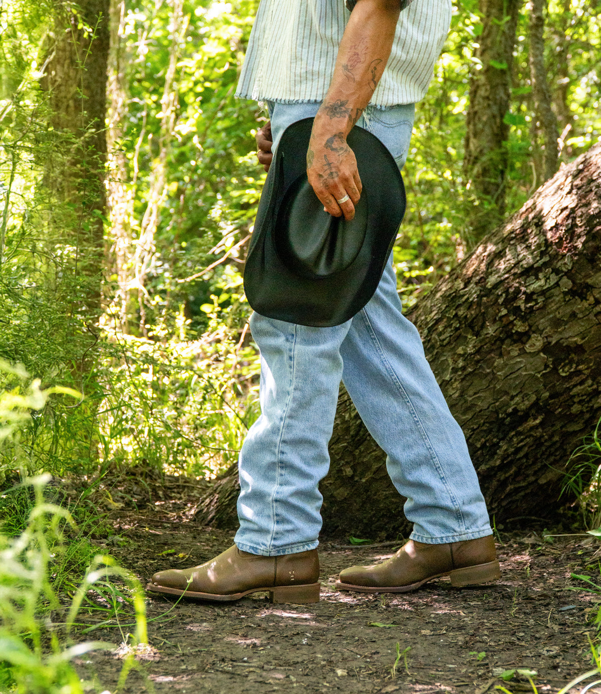 A person stands in a sunlit forest, showcasing Roan’s Satire boots with enhanced traction, paired with light blue jeans and a short-sleeve shirt. Only their lower half is visible, emphasizing rugged style and quality footwear.