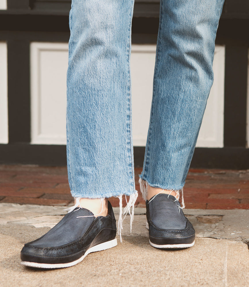 Close-up of a person wearing frayed-hem blue jeans and Roan Shevon dark-colored slip-on shoes with a canvas and leather upper, standing on a stone pavement.