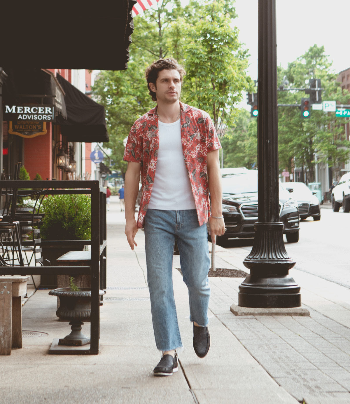 
                  
                    A man in a floral print shirt and jeans, sporting Roan Shevon leather slip-ons with cushioned insoles, walks on a city sidewalk near parked cars and buildings.
                  
                