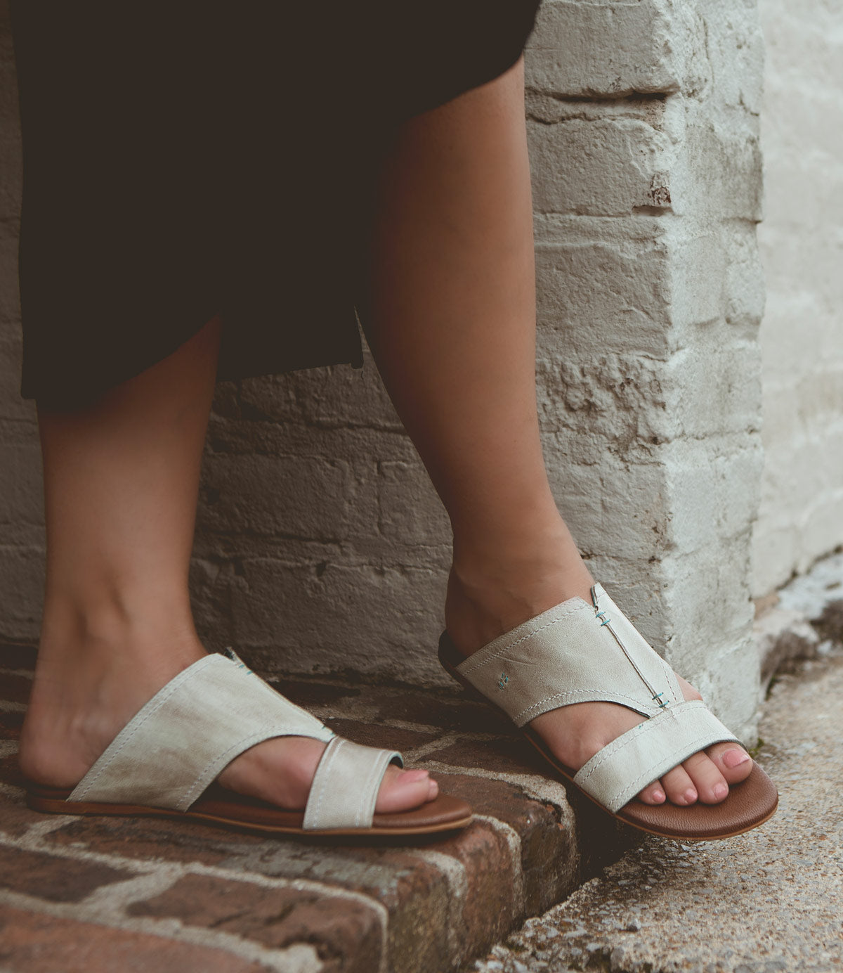 
                  
                    Person wearing all-leather, light-colored Roan Somerville summer slide sandals with a split toe design, standing on a brick surface near a white brick wall.
                  
                