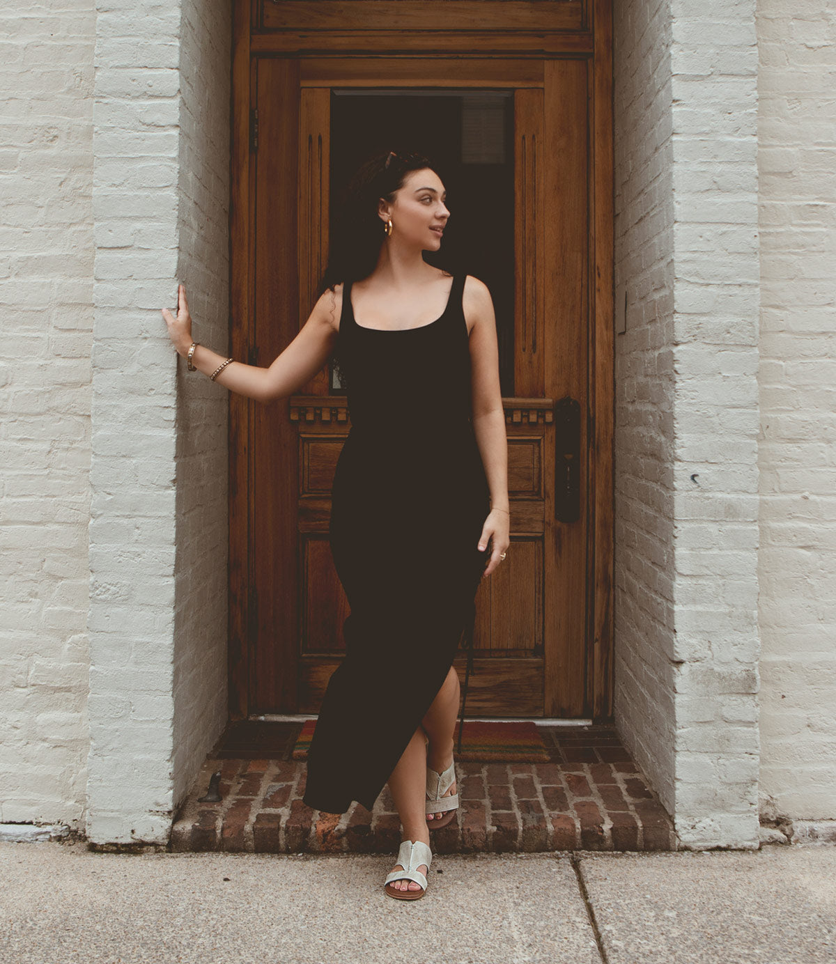 
                  
                    A woman in a black dress and Roan Somerville white leather slide sandals with a cushioned footbed stands in a doorway, touching the brick wall with her left hand.
                  
                