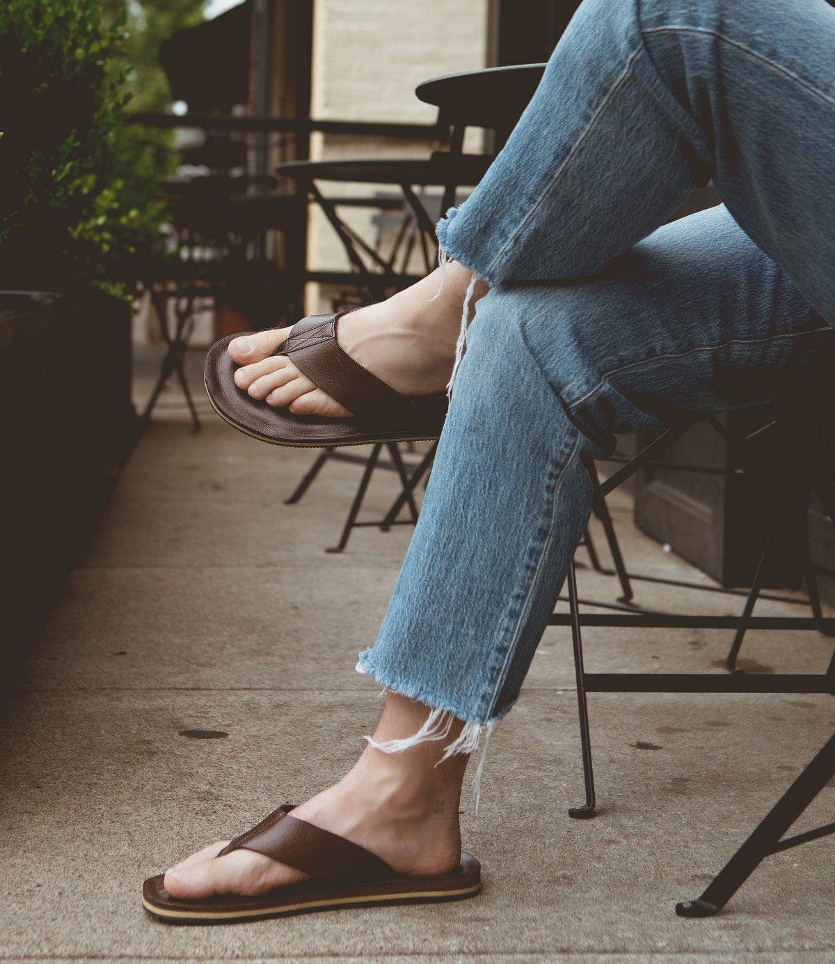 A person wearing Townfolk jeans and distressed leather sandals by Roan is seated outdoors with legs crossed on a patio with metal chairs and tables, adding a touch of rustic charm.