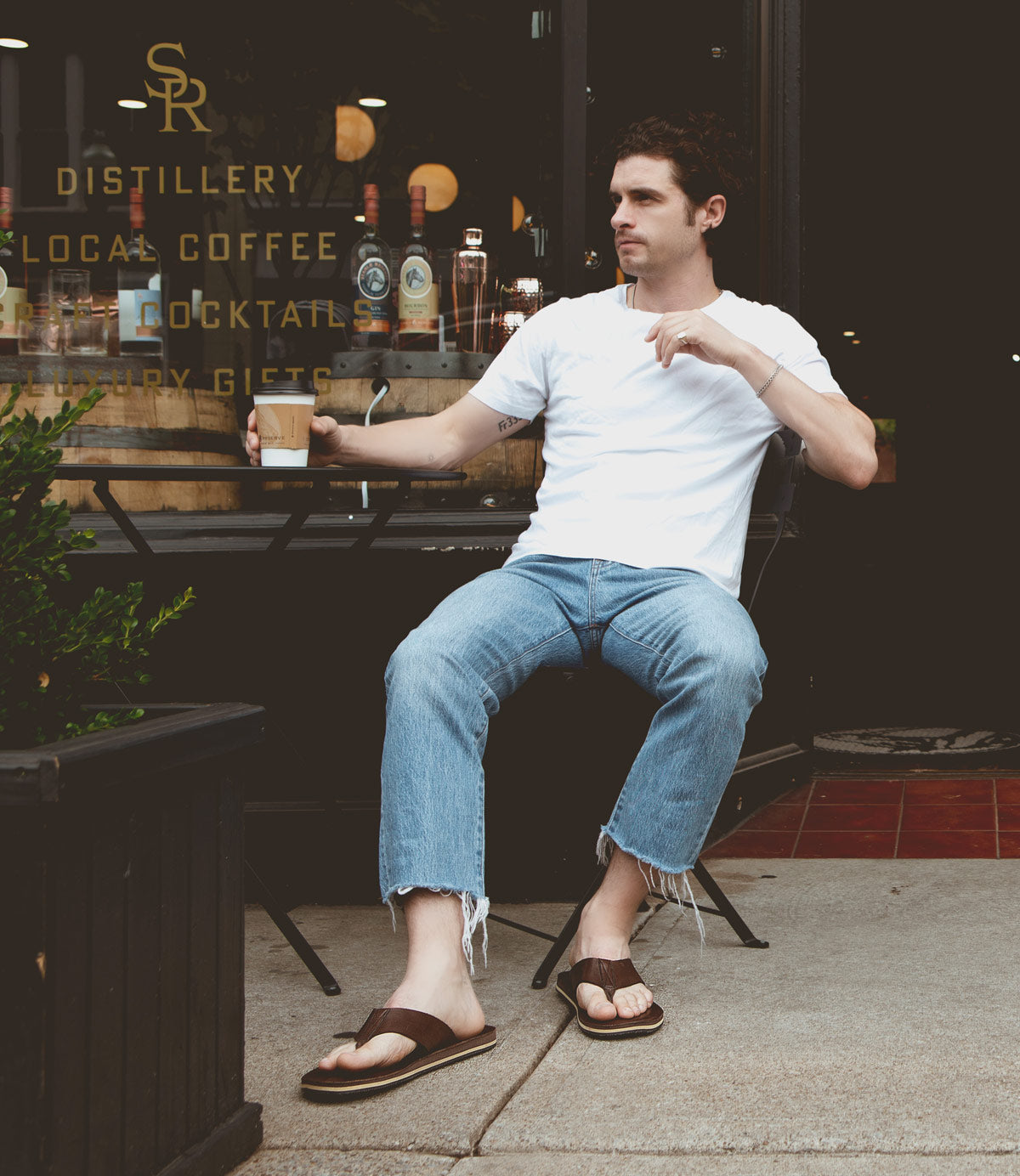 
                  
                    A man in a white t-shirt and jeans sits outside a cafe, holding a Townfolk by Roan in one hand. Wearing flip-flops and looking to his right, he exudes rustic charm against the backdrop of the cafe window's reflections and signage.
                  
                