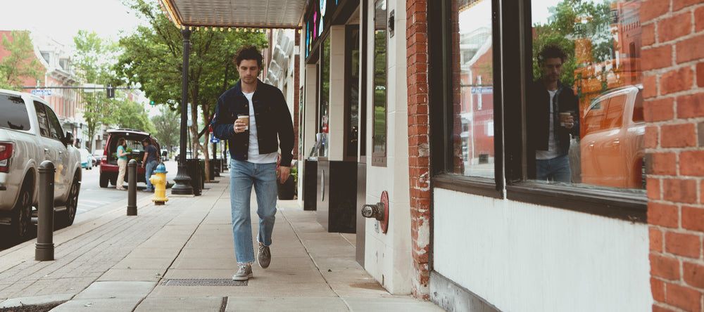 A person in casual attire walks down a city sidewalk holding a drink, with a brick building on the right and parked vehicles along the street.