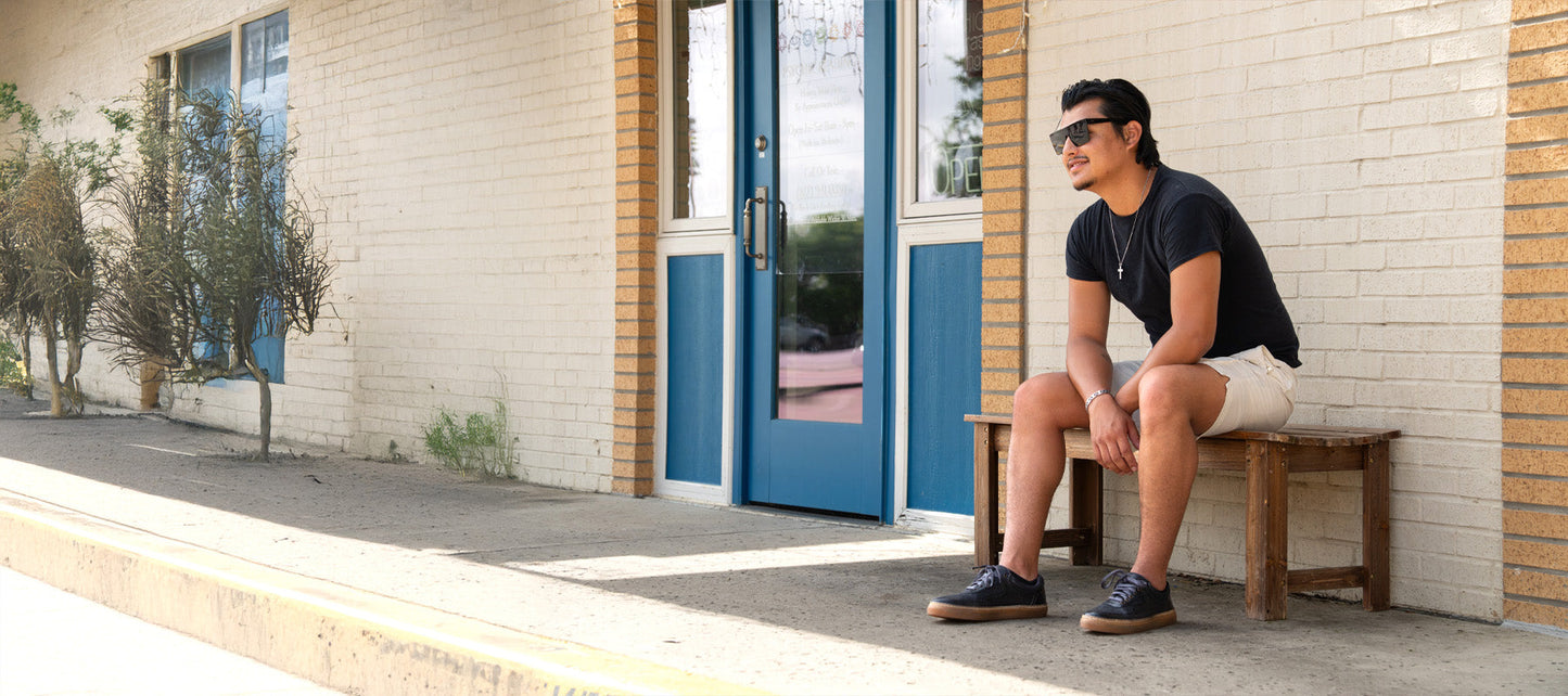 A man wearing sunglasses, a black t-shirt, and shorts sits on a wooden bench outside a building with blue doors and windows.