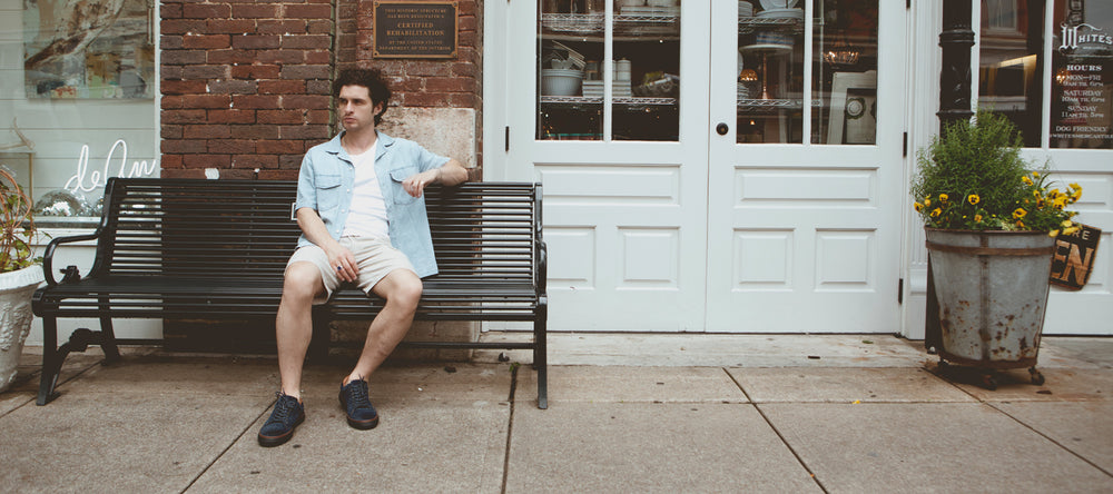 A man in casual clothing sits on a bench outside a brick building with white double doors and a window display.