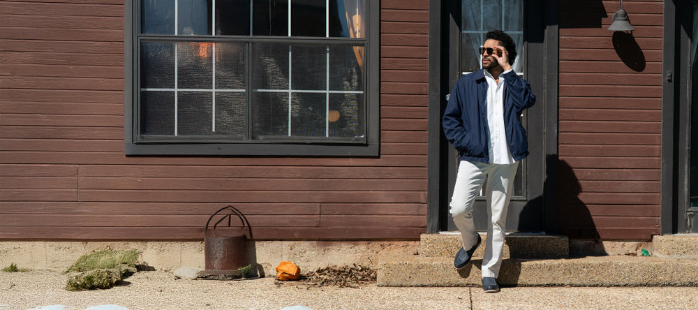 A man in a brown shirt and jeans stands outdoors on a dirt and wood-chipped area, wearing brown shoes.