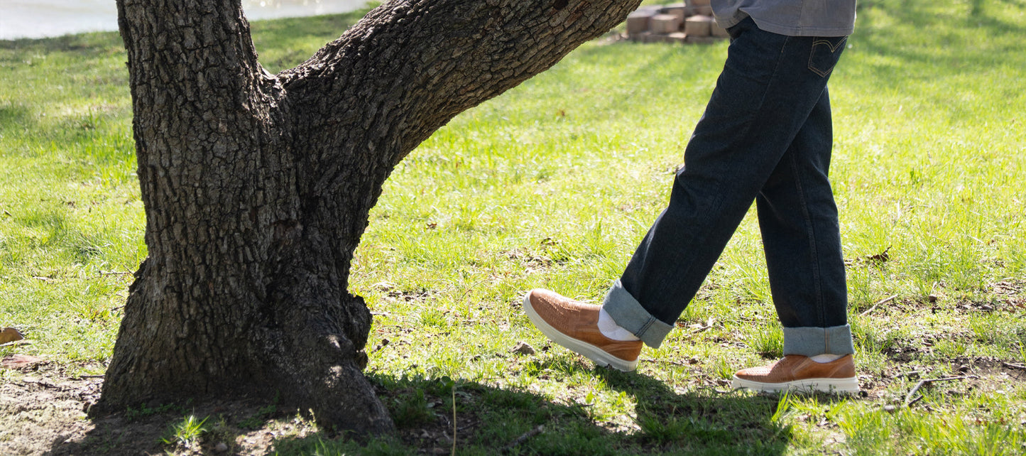 Person wearing dark jeans and tan shoes walks on grass near a tree in a sunlit outdoor setting.