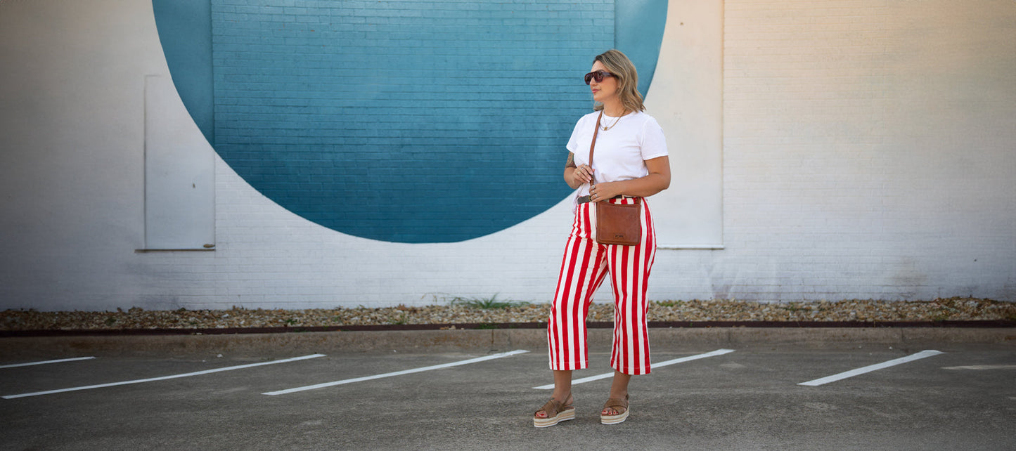 A person stands in a parking lot wearing a white t-shirt, red and white striped pants, sunglasses, and sandals, with a brown crossbody bag, in front of a wall with a large blue circle.