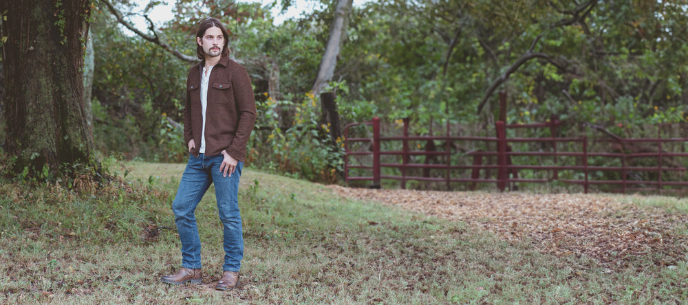 A man in casual attire standing in a grassy field with trees and a fence in the background.