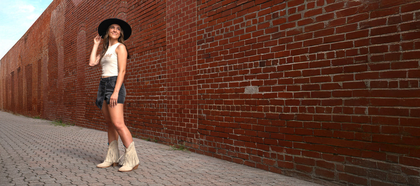 A woman wearing a black hat, white top, black shorts, and fringed boots stands on a brick sidewalk beside a long red brick wall.