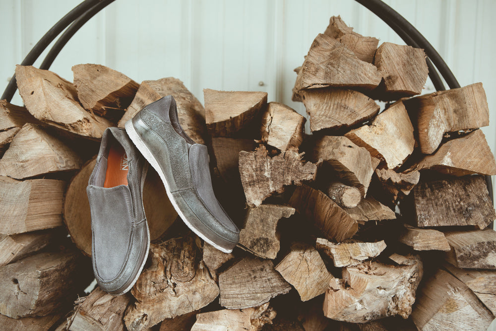 A pair of gray slip-on shoes placed on top of a pile of firewood against a wooden wall.
