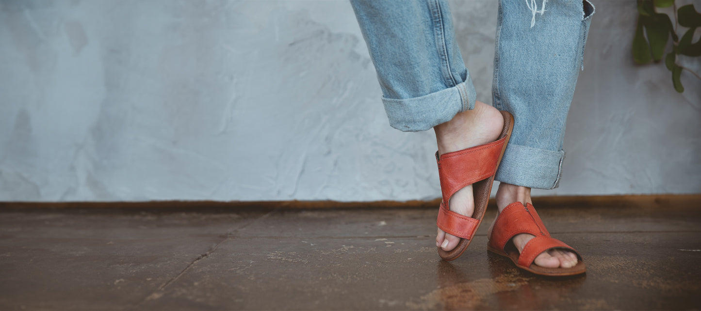 Close-up of a person's feet wearing red leather sandals and distressed blue jeans, standing on a textured brown floor against a grey wall.
