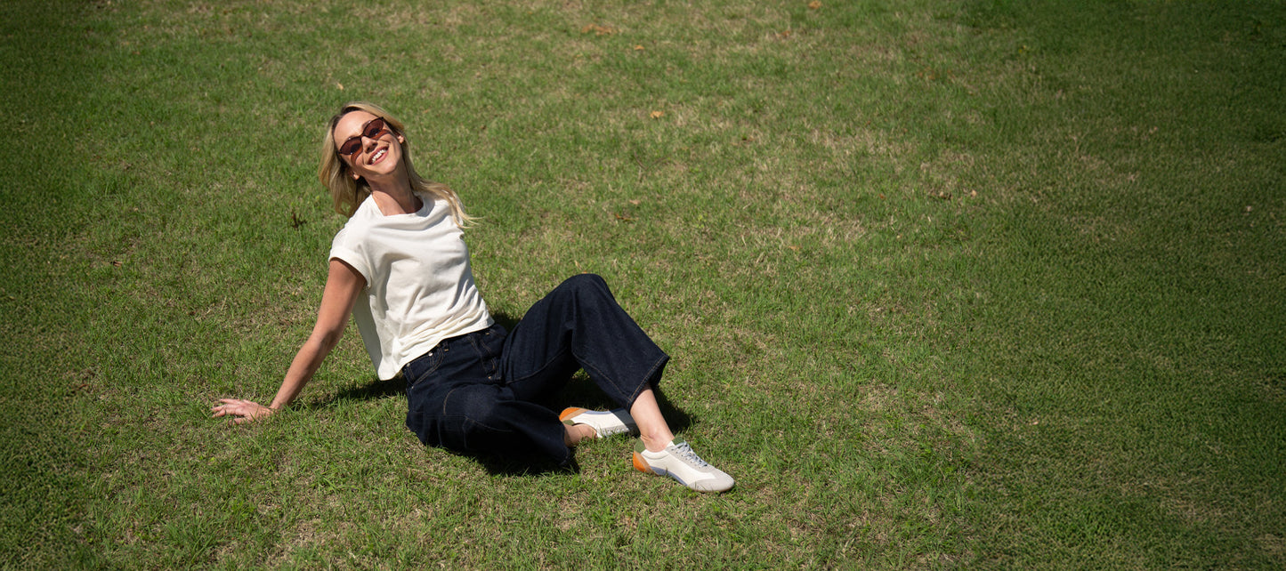 A person wearing sunglasses, a white t-shirt, dark pants, and white shoes sits on green grass, smiling and looking up.
