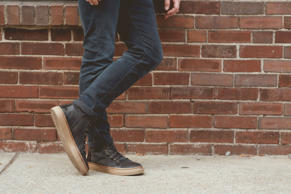 A person in dark jeans and black sneakers with tan soles leans against a brick wall.