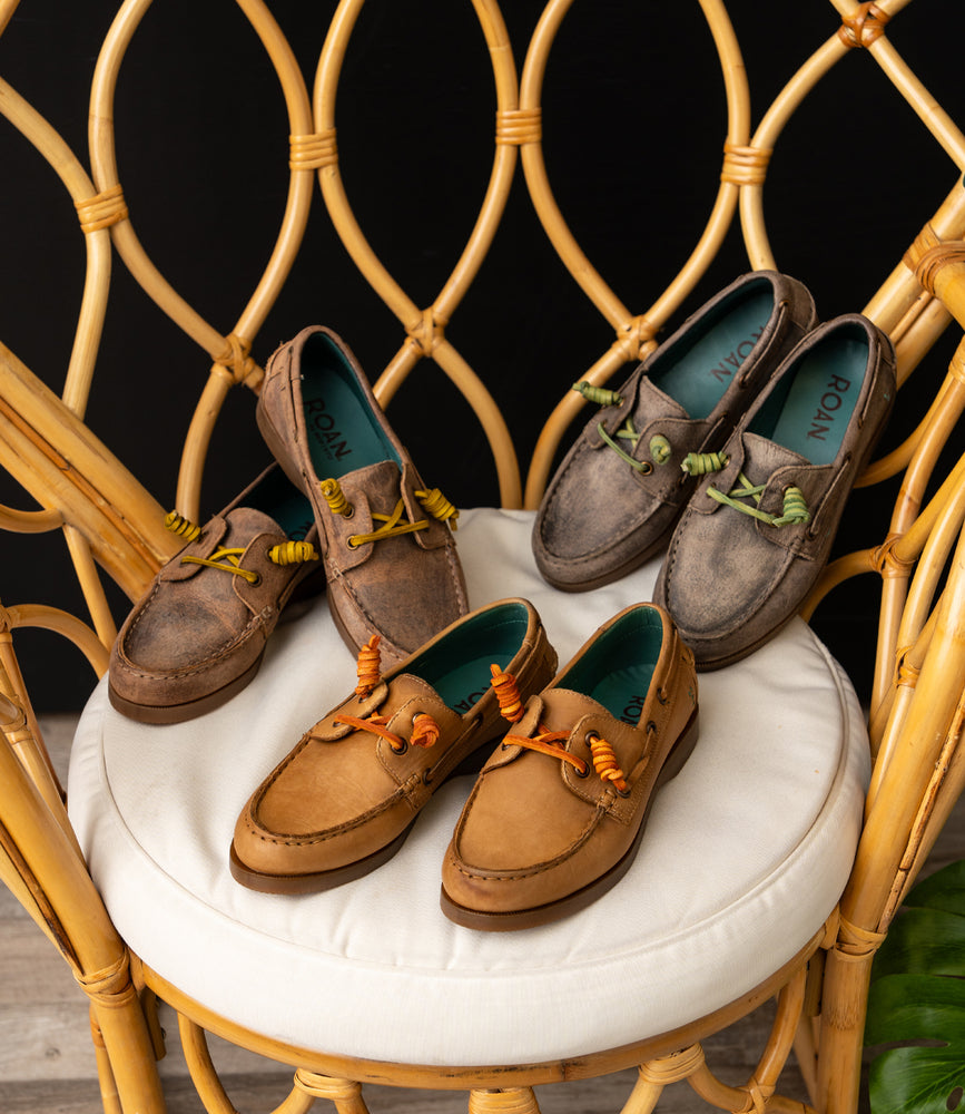 
                  
                    Four pairs of Roan Abide women's leather boat shoes in tan and gray, each with colored laces, are displayed on a cushioned rattan chair, highlighting their timeless style and versatile all-weather design.
                  
                
